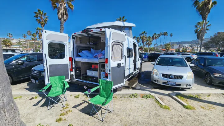 Relaxing day with an RV rental camper van featuring an open rear, bed, and chairs, parked by palm trees.. Winnebago Solis 2023