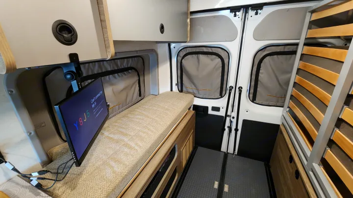 Modern RV rental interior showing a compact camper van layout with a bench seat, a monitor, and a slatted bed frame.. Winnebago Solis 2023
