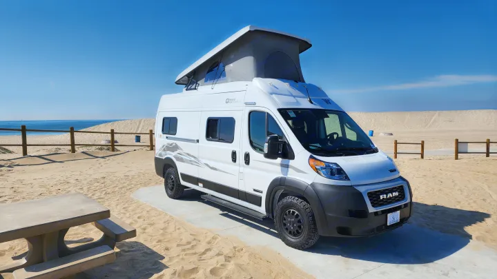 White pop-top RV rental parked at a sunny sandy beach with ocean views. Perfect for adventure!. Winnebago Solis 2023