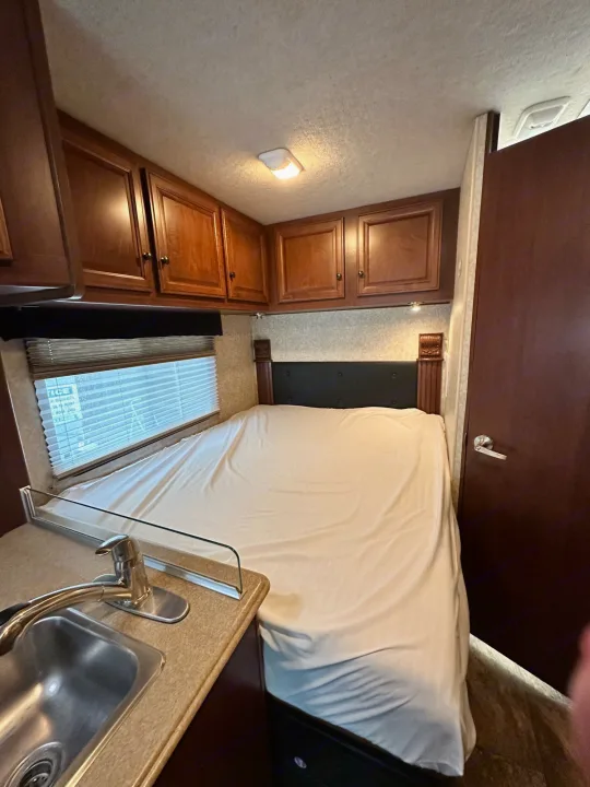 Inside this RV rental, a compact bedroom features a bed, overhead wood cabinets, and a kitchenette sink. Cozy interior for travel.. Jamboree Searcher 2013