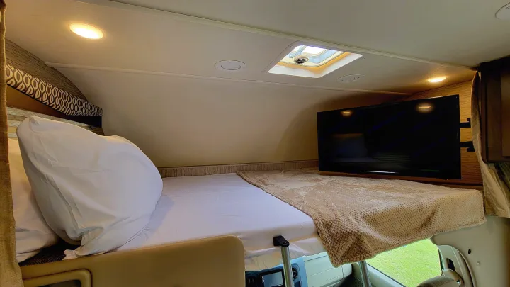 Cozy over-cab bunk bed in an RV rental, featuring a comfortable bed, TV, and bright lighting for restful trips.. Thor Freedom Elite 2021