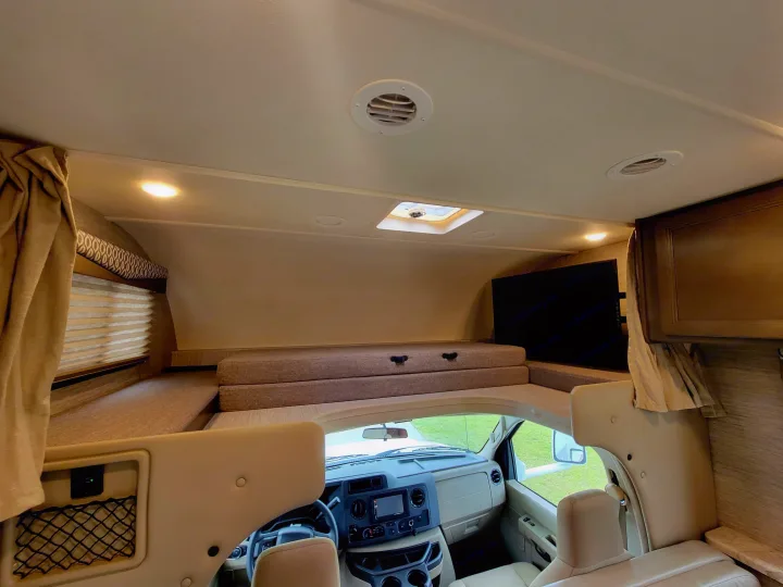 Inside this modern RV rental, find a cozy over-cab bunk bed, large TV, and the detailed front dashboard visible through the living space.. Thor Freedom Elite 2021