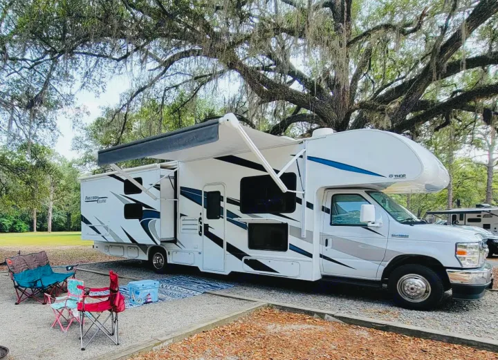 Spacious RV rental parked under mossy oak trees at a gravel campsite. Awning and outdoor seating create a cozy outdoor setup.. Thor Freedom Elite 2021