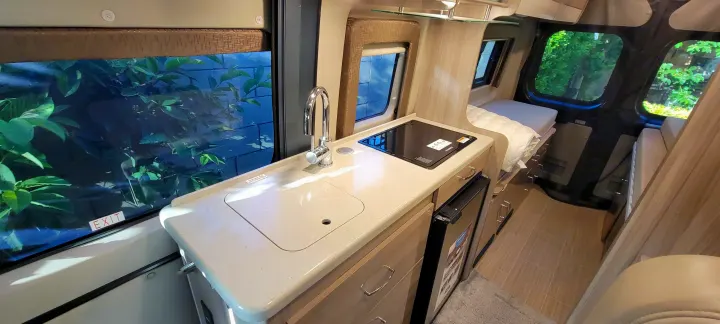 Bright RV rental interior showing a galley kitchen with sink, cooktop, fridge, and a built-in sleeping area.. Thor Sequence 2021