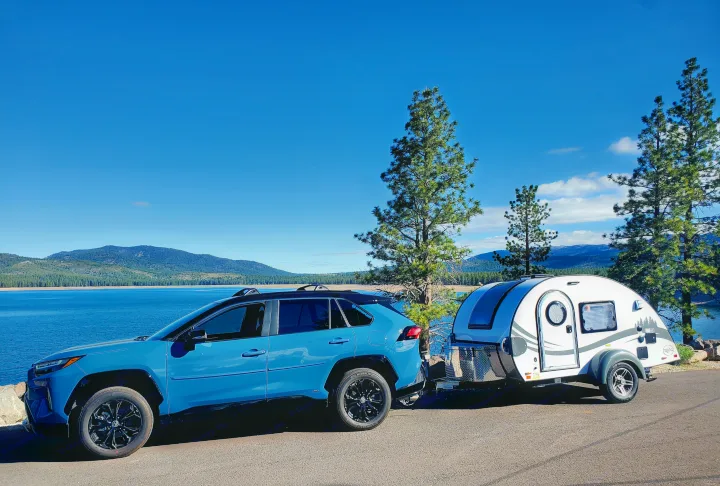A blue SUV with an RV rental teardrop trailer parked by a beautiful lake & mountains under a clear sky.. nuCamp T@G 2022