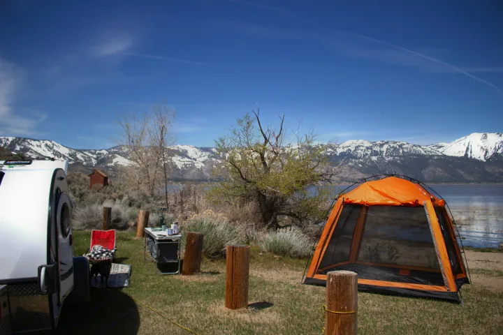 Lakeside campsite with a white RV rental, orange tent, and snow-capped mountains. Ideal for scenic nature camping.. nuCamp T@G 2022