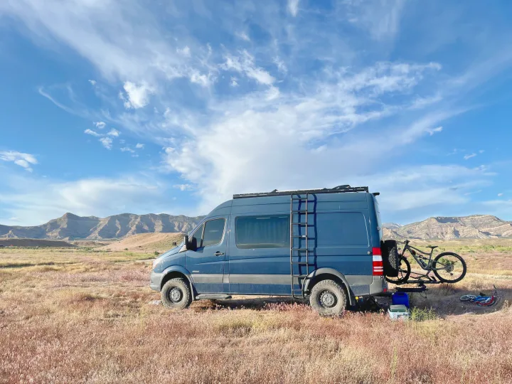 Dark blue RV rental van with mountain bike, parked in a vast field with mountains and a partly cloudy sky.. Mercedes Sprinter 2016
