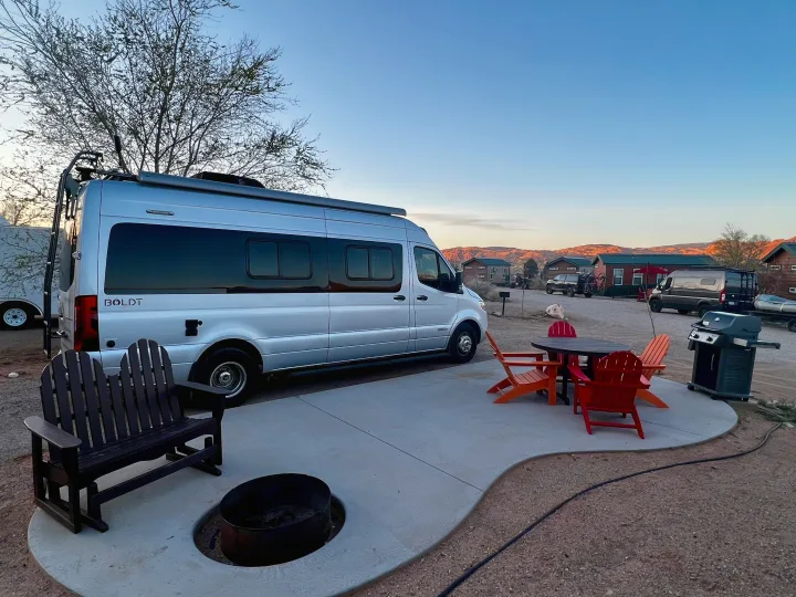 Silver Boldt campervan RV rental setup at a scenic campsite, complete with a patio, fire pit, and BBQ grill.. Winnebago Boldt 2020