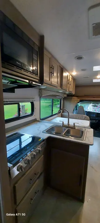 Spacious RV rental kitchen featuring a microwave, double sink, and stovetop. Light cabinets and windows show greenery.. Forest River Coachmen Freelander 2021