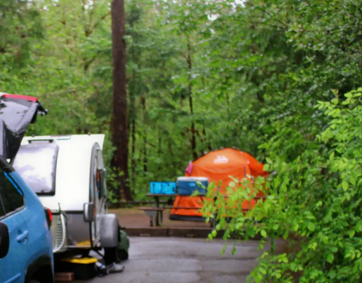 Campsite scene: A blue car and white teardrop RV rental are parked next to an orange tent, surrounded by lush green trees.. nuCamp T@G 2022