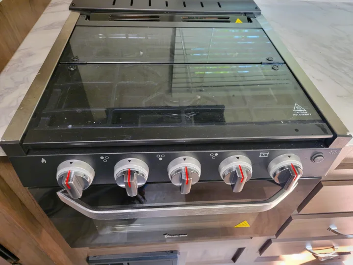 RV rental kitchen: compact black & silver stove and oven with glass top, control knobs, and oven handle, ready for cooking.. East to West Della Terra 2021