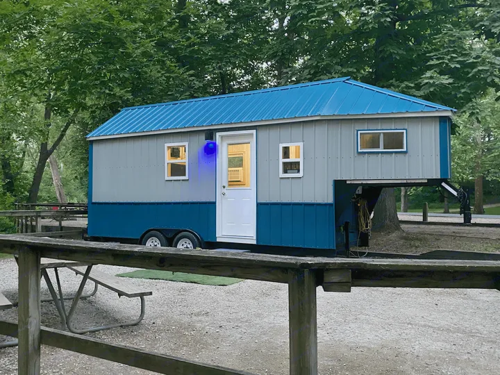This charming tiny home RV rental on wheels, with a blue roof, sits in a wooded campsite with a picnic table.. Bran Gooseneck 2019