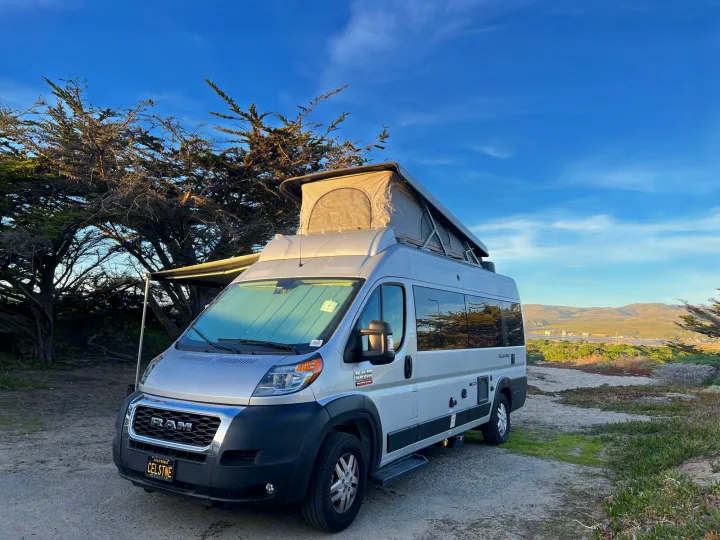 A silver Ram ProMaster pop-top camper van, possibly an rv rental, parked by coastal trees with a scenic mountain view.. Thor Tellaro 2021