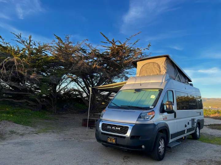 Experience RV rental adventures! Gray pop-top camper van at a scenic coastal campsite nestled by cypress trees.. Thor Tellaro 2021