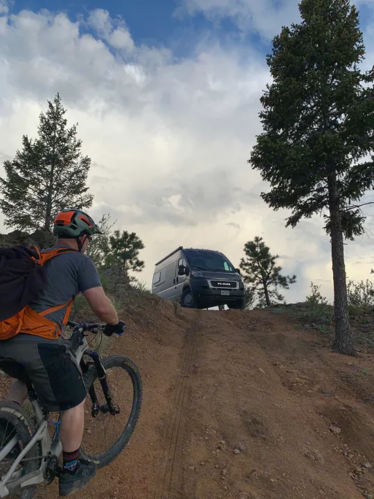 Outdoor adventure: A mountain biker heads up a dirt path towards a waiting RV rental van on a hilltop under a cloudy sky.. Dodge Ram Promaster 2500 2022