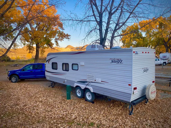 An RV rental, a white travel trailer and blue pickup truck, are set up at an autumn campsite with fallen leaves.. Jayco Jay Flight Swift 2013