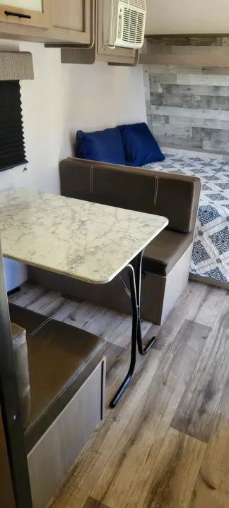 Inside an RV rental: a modern dinette with a marble-look table, bed, and AC unit on wood-patterned floor.. Dutchmen Coleman Lantern LT 2023