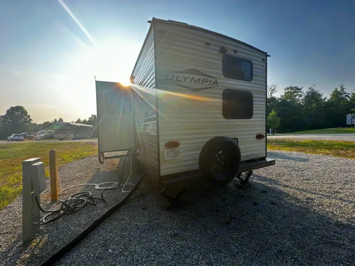 Bright sun flares behind an Olympia travel trailer, a potential rv rental, parked with hookups at a gravel campground site.. Highland Ridge RV Olympia 2022