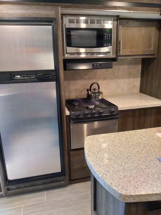 Modern kitchen in an RV rental, with stainless steel fridge, microwave, gas stove, and speckled light countertops.. Highland Ridge RV Open Range Ultra Lite 2018