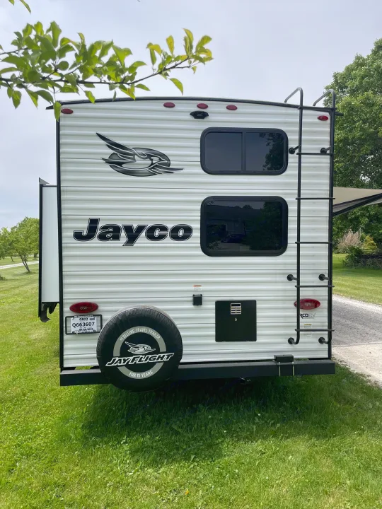 Rear view of a white Jayco travel trailer parked on grass, ideal for an RV rental. Features include a ladder & spare tire.. Jayco Jay Flight 2023