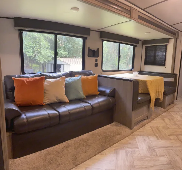 Inside a comfy RV rental: brown leather couch with colorful pillows, large windows, and a dinette. Carpet meets tile.. Keystone RV Springdale 2021