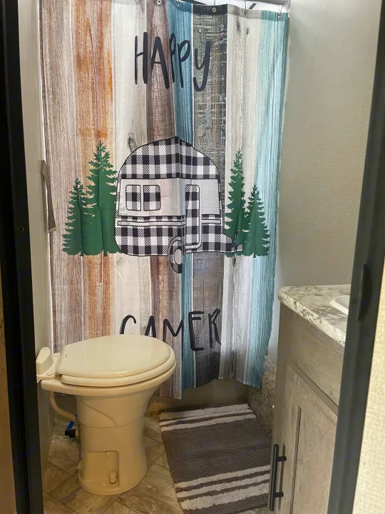 Compact RV rental bathroom with a toilet, sink area, and a themed "Happy Camper" shower curtain.. Keystone RV Springdale 2021