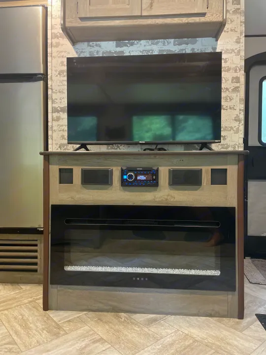Modern RV rental living area: A TV, Jensen stereo, & electric fireplace. White brick accent wall & fridge nearby.. Keystone RV Springdale 2021