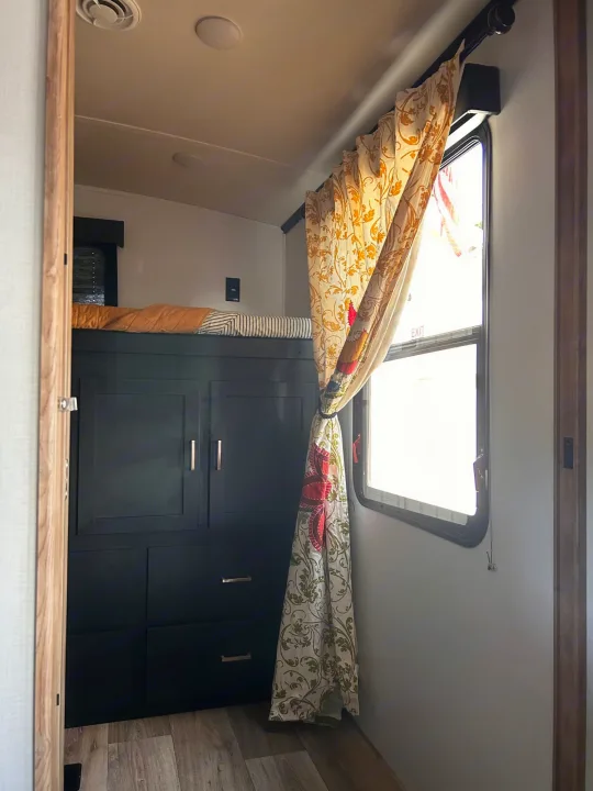 Cozy RV rental interior showing bunk beds, black storage cabinets, and colorful tied-back curtains by a window.. Keystone RV Outback Ultra-Lite 2022