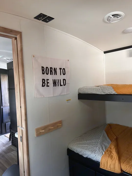 Modern RV rental bunk room with "Born To Be Wild" flag, wooden pegs, and cozy striped beds.. Keystone RV Outback Ultra-Lite 2022
