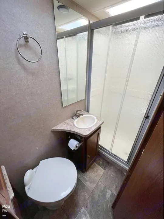 This clean RV rental bathroom features a compact toilet, sink vanity with mirror, and a frosted glass shower stall.. Coachmen Leprechaun 2015