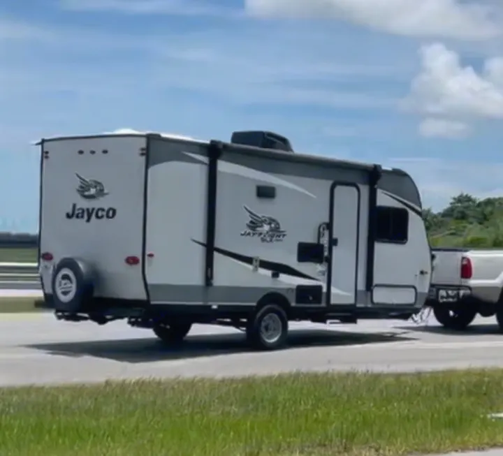 White & gray Jayco travel trailer RV rental, with a spare tire on back, towed by a white pickup truck on a road.. Jayco Jay Flight 2021