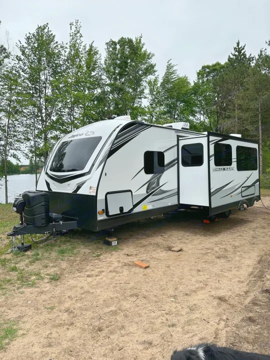 White Jayco RV rental at a lakeside campsite with trees. Slide-out extended for outdoor comfort.. Jayco Whitehawk 2023