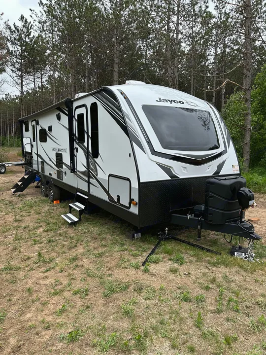 A modern Jayco travel trailer, ready for an RV rental adventure, is nestled in a rustic, tree-lined campsite. Steps are down.. Jayco Whitehawk 2023