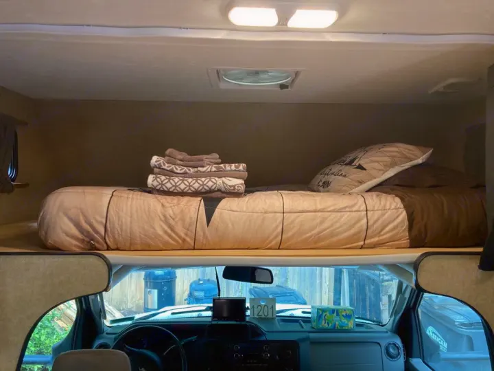 Inside an RV rental, a comfy overhead bed with bedding & towels, seen from the driver's cab.. Thor Majestic 2016