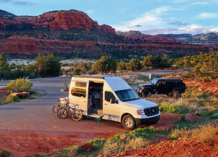 White RV rental camper van with bikes parked roadside, with striking red rock mountains in background. A black Jeep is also visible.. Nissan Other 2012