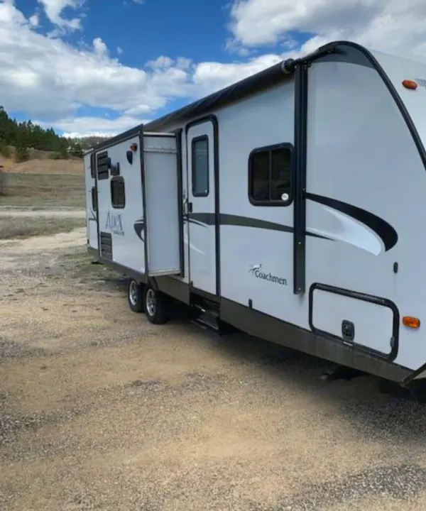 A white Coachmen travel trailer RV rental, with its side door open, is parked on gravel under a blue sky.. Coachmen Apex 2015
