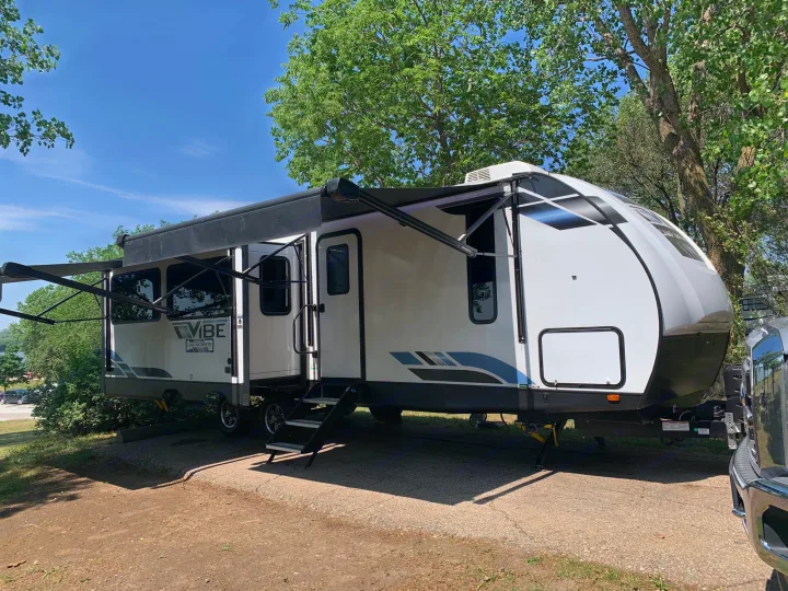 White VIBE travel trailer parked at a tree-lined campsite with awnings extended, ideal for an RV rental getaway.. Forest River Vibe 2023