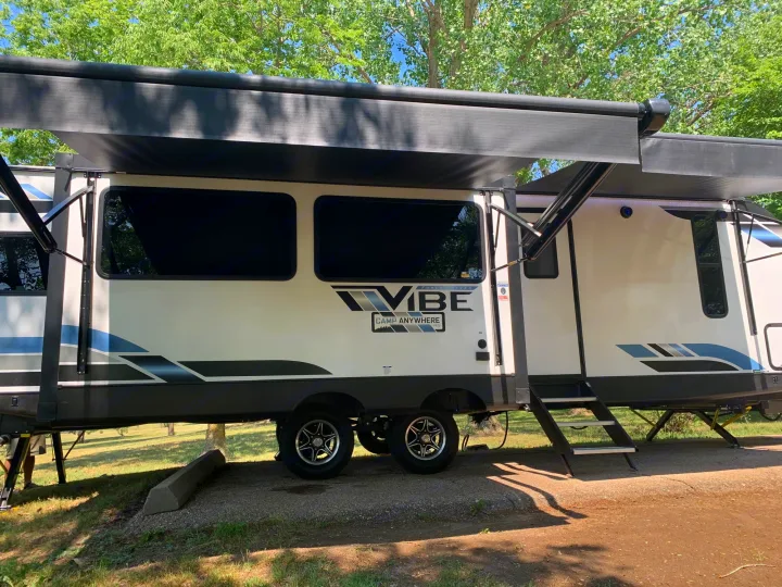 A white Forest River Vibe travel trailer, likely an RV rental, with awnings extended at a sunny campsite.. Forest River Vibe 2023