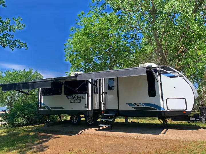 White and grey Vibe travel trailer RV rental with awning extended, parked in a scenic campground by water.. Forest River Vibe 2023