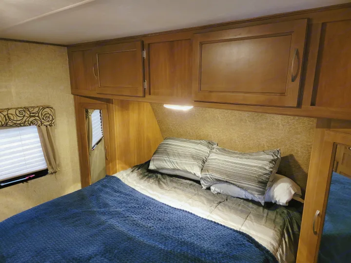 Cozy RV rental bedroom with wood cabinets, comfy bed, and window, offering a compact, inviting interior space.. Coachmen Apex 2015