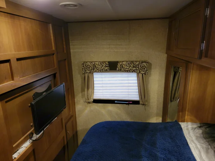Inside an RV rental: a compact bedroom featuring a cozy bed, window with blinds, and wood cabinetry with a mounted TV.. Coachmen Apex 2015