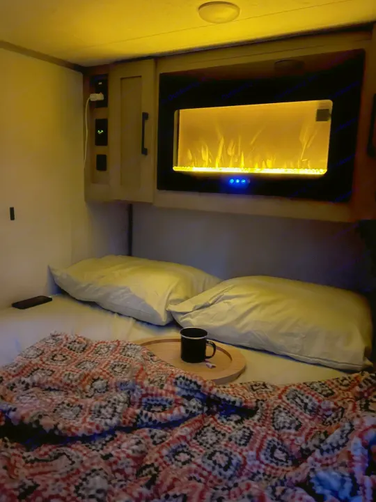 Cozy RV rental bedroom features an electric fireplace, soft pillows, a warm blanket, and a mug on a tray for comfort.. Dutchmen Coleman Rubicon 2023