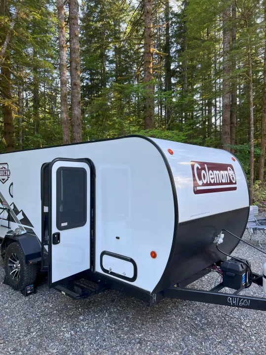 White Coleman travel trailer RV rental parked on gravel in a dense forest. Steps extended, ready for camping.. Dutchmen Coleman Rubicon 2023