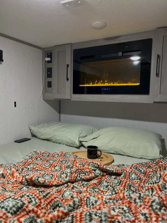 Cozy RV rental bedroom with electric fireplace, bed, light green pillows, patterned blanket, and tea.. Dutchmen Coleman Rubicon 2023