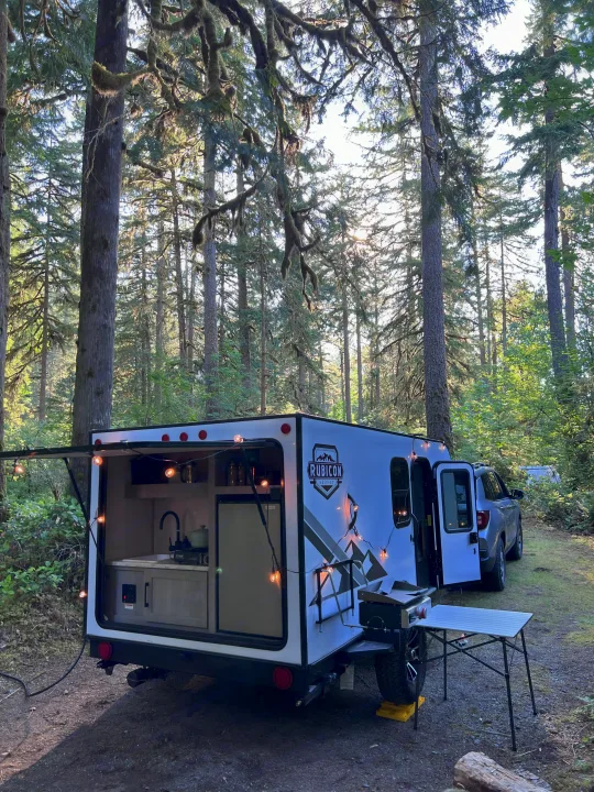 An RV rental in a lush forest, featuring a white teardrop camper with an open outdoor kitchen and string lights.. Dutchmen Coleman Rubicon 2023