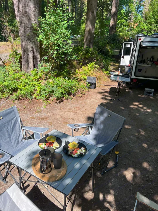 RV rental camping breakfast in a sunny forest. Table with plates of food, French press & chairs create a cozy setup.. Dutchmen Coleman Rubicon 2023