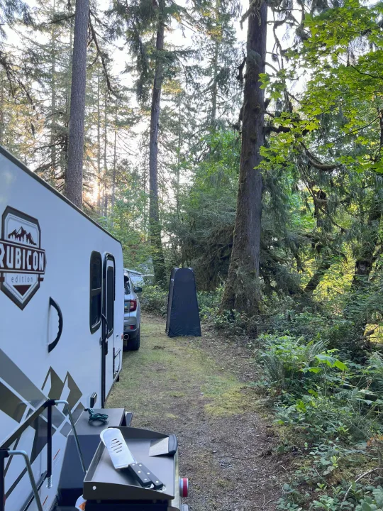 RV rental camping: A white Rubicon travel trailer is set up in a beautiful, mossy forest campsite with a black privacy tent.. Dutchmen Coleman Rubicon 2023