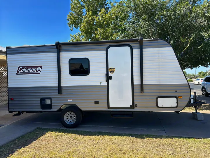 This Coleman travel trailer, ideal for an rv rental, is parked on a sunny day. A gray and white camper with a door & window.. Coleman Lantern LT 17B 2023