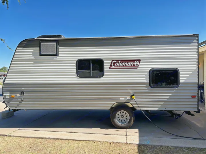 A Coleman travel trailer RV rental, light grey with corrugated siding, is parked on pavement hooked to power.. Coleman Lantern LT 17B 2023