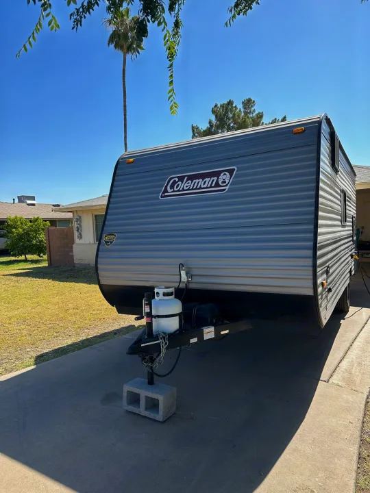 A gray Coleman travel trailer (RV rental) parked on a sunny driveway. Propane tanks visible at the front hitch.. Coleman Lantern LT 17B 2023